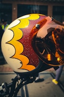 A close-up view of a helmet with a retro-style design featuring yellow and red scalloped patterns on a cream background. The helmet has an orange-tinted visor that reflects urban surroundings. The background is slightly blurred, showing a street scene with a few indistinct figures.