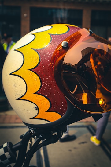 A close-up view of a helmet with a retro-style design featuring yellow and red scalloped patterns on a cream background. The helmet has an orange-tinted visor that reflects urban surroundings. The background is slightly blurred, showing a street scene with a few indistinct figures.