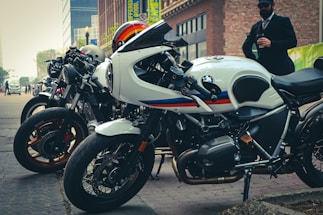 A row of sleek motorcycles is parked on a city street. The foreground features a prominently visible white BMW bike with a sporty design. Several other motorcycles line up alongside it, showcasing various designs and colors. In the background, there are tall buildings, and a person dressed in formal attire stands nearby holding a phone.