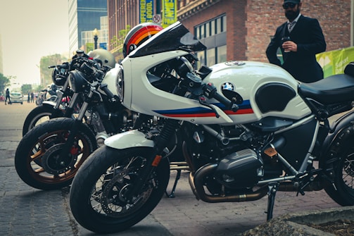 A row of sleek motorcycles is parked on a city street. The foreground features a prominently visible white BMW bike with a sporty design. Several other motorcycles line up alongside it, showcasing various designs and colors. In the background, there are tall buildings, and a person dressed in formal attire stands nearby holding a phone.