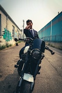 A person wearing a suit sits on a motorcycle parked in an urban alley. Their hand is extended, pointing towards the camera. On one side, there is a building with graffiti art, and on the other side, a blue industrial structure with a chain-link fence.