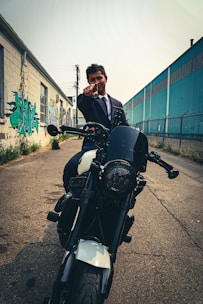A person wearing a suit sits on a motorcycle parked in an urban alley. Their hand is extended, pointing towards the camera. On one side, there is a building with graffiti art, and on the other side, a blue industrial structure with a chain-link fence.
