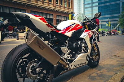 A sleek, white and red sports motorcycle is parked on a city street. The motorcycle is prominently branded with 'Honda' and features a large Akrapovic exhaust. In the background, there are modern buildings and other motorcycles, as well as a person wearing a red checkered shirt.