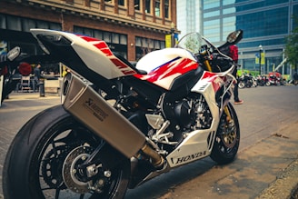 A sleek, white and red sports motorcycle is parked on a city street. The motorcycle is prominently branded with 'Honda' and features a large Akrapovic exhaust. In the background, there are modern buildings and other motorcycles, as well as a person wearing a red checkered shirt.