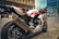 A sleek, white and red sports motorcycle is parked on a city street. The motorcycle is prominently branded with 'Honda' and features a large Akrapovic exhaust. In the background, there are modern buildings and other motorcycles, as well as a person wearing a red checkered shirt.