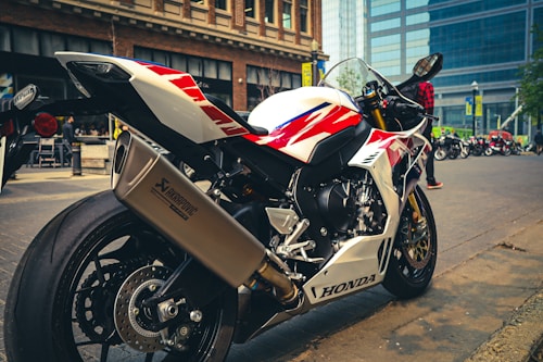 A sleek, white and red sports motorcycle is parked on a city street. The motorcycle is prominently branded with 'Honda' and features a large Akrapovic exhaust. In the background, there are modern buildings and other motorcycles, as well as a person wearing a red checkered shirt.