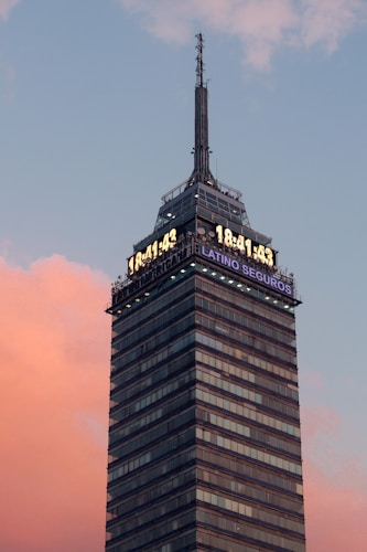A tall, modern skyscraper with a digital clock display near the top, showing the time 18:41:43. The building has many windows and an antenna at the very top. The sky in the background is a gradient of blue and pink, suggesting either dawn or dusk.