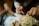 Hands are applying makeup around the eye area. The detailed view focuses on the precision of the application, with attention to lashes and eyeshadow. The subject is wearing an ornate, gold headpiece with intricate detailing.