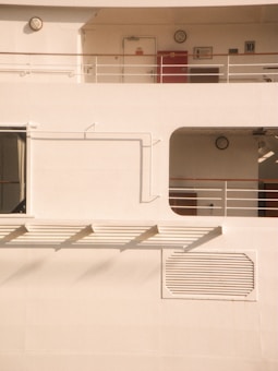 A minimalist view of a ship's exterior with clean lines and geometric shapes. There are several rectangular sections, including a door, a red panel, and a vent. The structure features horizontal railings and portholes. The lighting creates soft shadows on the cream-colored surface.