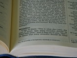 A close-up view of an open book displaying printed text in two columns, with a particular focus on the words 'Wineskins' and a passage discussing the commands of the Lord. The pages are slightly curved, indicating the book is either new or gently opened. The light casts a soft shadow, adding depth to the image.
