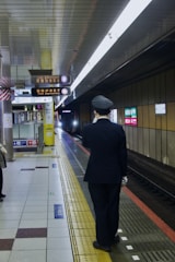 A train station platform with a person wearing a uniform, standing close to the edge. The platform has signage indicating an approaching train, and the lighting is bright, illuminating the tiled floor. Advertising signs and informational indicators are visible on the walls.