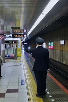 A train station platform with a person wearing a uniform, standing close to the edge. The platform has signage indicating an approaching train, and the lighting is bright, illuminating the tiled floor. Advertising signs and informational indicators are visible on the walls.