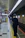 A train station platform with a person wearing a uniform, standing close to the edge. The platform has signage indicating an approaching train, and the lighting is bright, illuminating the tiled floor. Advertising signs and informational indicators are visible on the walls.