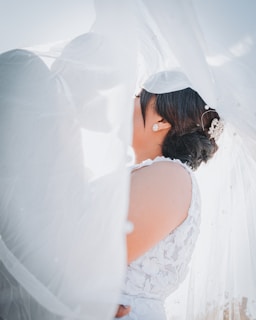 A bride in a white floral-patterned gown is partially obscured by a flowing veil. The veil creates a soft, dreamy effect as it drapes over the scene, highlighting the bride's intricate hairstyle and decorative hairpiece. Bright sunlight filters through, creating a warm and ethereal atmosphere.