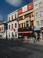 A street scene featuring several buildings with signage in Turkish. The architecture is a mix of traditional and modern styles, with banners displaying the Turkish flag. People walk along the sidewalk, some entering and exiting storefronts. The sky is clear with a few clouds, and tram tracks run along the road.
