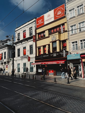 A street scene featuring several buildings with signage in Turkish. The architecture is a mix of traditional and modern styles, with banners displaying the Turkish flag. People walk along the sidewalk, some entering and exiting storefronts. The sky is clear with a few clouds, and tram tracks run along the road.