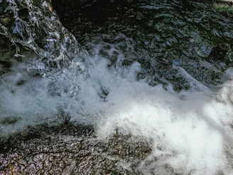 A clear and vibrant stream of water cascades down, creating frothy bubbles and splashes as it interacts with darker, textured water below. The dynamic movement and play of light capture the mesmerizing flow and energy of the water.