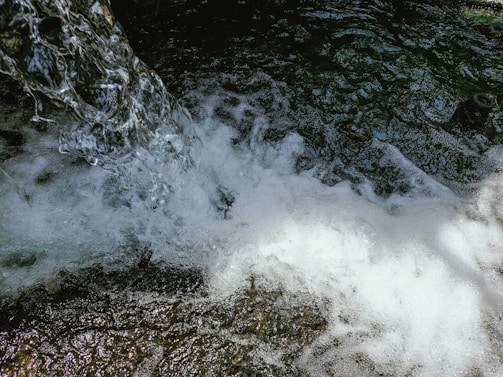 A clear and vibrant stream of water cascades down, creating frothy bubbles and splashes as it interacts with darker, textured water below. The dynamic movement and play of light capture the mesmerizing flow and energy of the water.