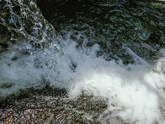 A clear and vibrant stream of water cascades down, creating frothy bubbles and splashes as it interacts with darker, textured water below. The dynamic movement and play of light capture the mesmerizing flow and energy of the water.