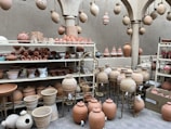 A collection of handcrafted pottery displayed on shelves and stands, including various shapes and sizes of pots, vases, and jugs. Hanging from arches are intricately designed lanterns. The setting has a rustic and traditional feel, with earthy tones and craftsman-style pillars.