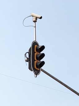 A traffic light mounted on a pole with a surveillance camera attached above it. The sky in the background is clear and blue.