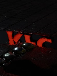 Dark pavement with a red reflection of letters 'KLC' in a puddle. Silver metallic orbs are positioned on the wet surface, adding a futuristic or abstract element to the scene.