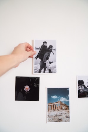 A hand is pinning a black and white photograph on a white wall. The photo features a playful scene where one person gives another a piggyback ride, and both are smiling. Around the main photo, there's an image of a pink flower against a dark background, a color photograph of an ancient Greek ruin under a blue sky, and another close-up black and white photo of a person smiling.