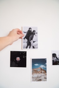 A hand is pinning a black and white photograph on a white wall. The photo features a playful scene where one person gives another a piggyback ride, and both are smiling. Around the main photo, there's an image of a pink flower against a dark background, a color photograph of an ancient Greek ruin under a blue sky, and another close-up black and white photo of a person smiling.