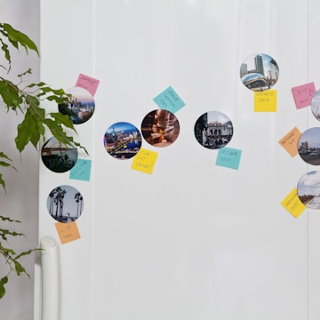 A refrigerator door is decorated with circular travel photos and colorful sticky notes. The images depict scenes from various locations, while the notes label each place with handwritten text. A green potted plant partially frames the left side of the image.