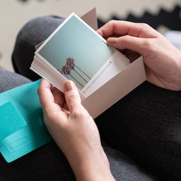 A person is holding a stack of square photographs, with the top photo showing a clear blue sky and palm trees. The person has manicured nails and is wearing dark pants. There is also an open envelope in a light brown color and a teal card visible in the scene.