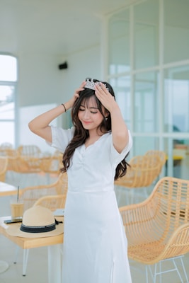 A young woman in a white dress is adjusting a tiara on her head. She is standing in a bright, modern cafe with wicker chairs and a wooden table. A straw hat, drink, and book are on the table next to her.
