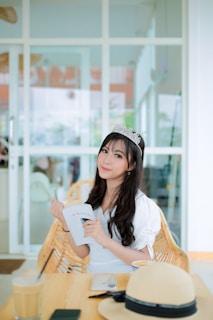 A woman wearing a tiara is seated at a wooden table holding a book. She is dressed in a white outfit and has long dark hair. The background features large glass windows and doors, allowing natural light to illuminate the room. On the table, there is a hat, a phone, and a glass of iced coffee.
