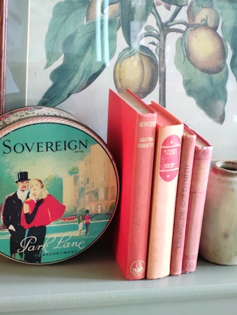A vintage illustration depicting a couple dressed in classic attire adorns a circular tin box labeled 'Park Lane Assortment.' Next to it, there are several hardcover books in shades of red and pink, including titles by Agatha Christie and Oliver Goldsmith. In the background, a framed botanical print featuring green leaves and yellow fruits is visible, adding a touch of nature to the setting. An earthenware vase stands to the right of the books.