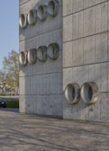 A modern architectural wall made of concrete panels featuring multiple circular protrusions arranged in a grid pattern. The smooth, industrial design gives it a minimalist aesthetic. A car is parked to the left, and trees are visible in the background, indicating an urban setting. The ground is paved with patterned stone tiles.