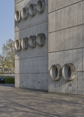 A modern architectural wall made of concrete panels featuring multiple circular protrusions arranged in a grid pattern. The smooth, industrial design gives it a minimalist aesthetic. A car is parked to the left, and trees are visible in the background, indicating an urban setting. The ground is paved with patterned stone tiles.