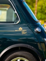 A close-up view of a classic car, featuring the rear window, wheel arch, and side panel, with a badge reading 'BRITISH OPEN Classic' in gold lettering. The car's paint is a deep green, and the chrome detailing is visible around the window.