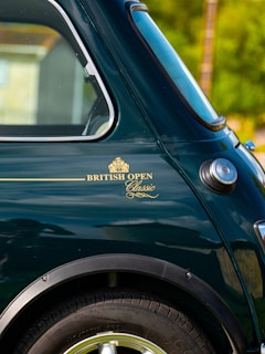 A close-up view of a classic car, featuring the rear window, wheel arch, and side panel, with a badge reading 'BRITISH OPEN Classic' in gold lettering. The car's paint is a deep green, and the chrome detailing is visible around the window.