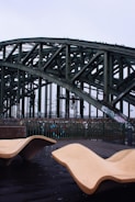 An industrial-style bridge with a lattice structure towers above a walkway. Modern, abstract benches made of a smooth light-colored material are positioned in the foreground, contrasting with the dark, wet surface of the ground.