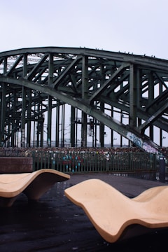 An industrial-style bridge with a lattice structure towers above a walkway. Modern, abstract benches made of a smooth light-colored material are positioned in the foreground, contrasting with the dark, wet surface of the ground.