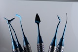 A set of six shiny blue metal instruments with various shaped ends, displayed vertically against a plain background. Each tool has a unique, intricate design, suggesting they are specialized for specific tasks.