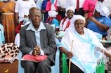 A group of people is gathered, with two elderly individuals seated at the forefront. One person wears a striped suit and sits with hands clasped, while the other is dressed in white and blue ceremonial attire, holding a walking stick. The background features individuals in colorful clothing and headscarves, some wearing masks.