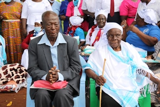 A group of people is gathered, with two elderly individuals seated at the forefront. One person wears a striped suit and sits with hands clasped, while the other is dressed in white and blue ceremonial attire, holding a walking stick. The background features individuals in colorful clothing and headscarves, some wearing masks.
