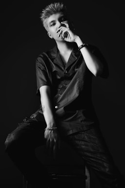 A person with short, styled hair sits on a stool against a dark background. They wear a dark button-up shirt and pants, with a watch on one wrist and a bracelet on the other. The lighting creates strong contrasts, casting shadows across the face and body.
