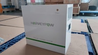 A white energy storage system labeled with 'HARVEYPOW' is situated in an industrial setting, surrounded by cardboard and wooden pallets. There is a green stripe on the unit with the words 'Energy storage system' beneath the brand name. The environment appears to be a warehouse.