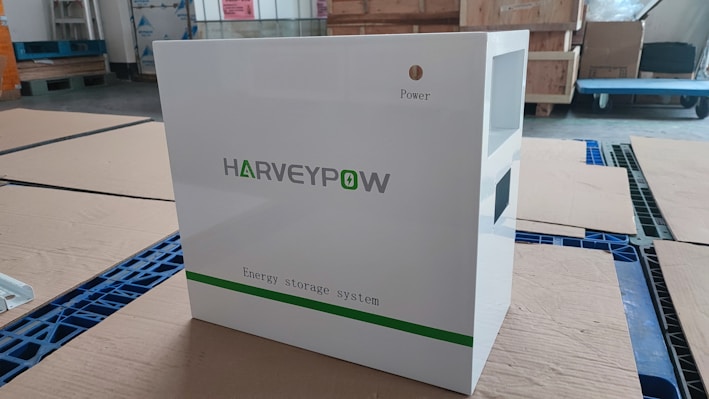 A white energy storage system labeled with 'HARVEYPOW' is situated in an industrial setting, surrounded by cardboard and wooden pallets. There is a green stripe on the unit with the words 'Energy storage system' beneath the brand name. The environment appears to be a warehouse.