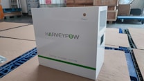 A white energy storage system labeled with 'HARVEYPOW' is situated in an industrial setting, surrounded by cardboard and wooden pallets. There is a green stripe on the unit with the words 'Energy storage system' beneath the brand name. The environment appears to be a warehouse.