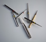A collection of dental or engraving drill bits arranged on a smooth, light surface. These tools vary in size and shape, with metallic bodies and different colored rings near their tips for identification. Some have diamond or grit-coated tips, while others have a smooth finish, and one has a white conical shape.