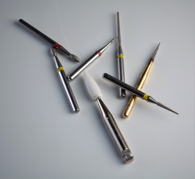 A collection of dental or engraving drill bits arranged on a smooth, light surface. These tools vary in size and shape, with metallic bodies and different colored rings near their tips for identification. Some have diamond or grit-coated tips, while others have a smooth finish, and one has a white conical shape.