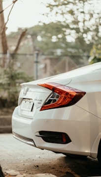 A white Honda car in a parking area with surrounding greenery and trees. The image focuses on the back of the vehicle, displaying its modern design, taillights, and branding details.