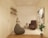 A cozy, minimalist room with neutral tones featuring a comfortable armchair, a bean bag, a small side table with plants, and decorative artwork on the walls. The shelves on the left hold various books and small decor items. The room has a modern white textured wall and wooden flooring with a geometric pattern rug.